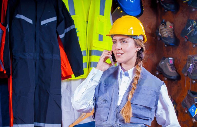 Frau mit Helm telefoniert, im Hintergrund Schutzkleidung und Sicherheitsausrüstung.